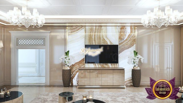 This picture shows a luxurious interior design in a modern office space. The walls are decorated with large, light-colored tiles, and the ceiling is adorned with hanging plants along its perimeter. A black leather sofa with gold accents is centered on the room and is surrounded by two black arm chairs, a round glass table and a large vase of fresh flowers. On the wall to the left is a collection of framed artwork and to the right is a modern fireplace with a checkered patterned floor rug below it.