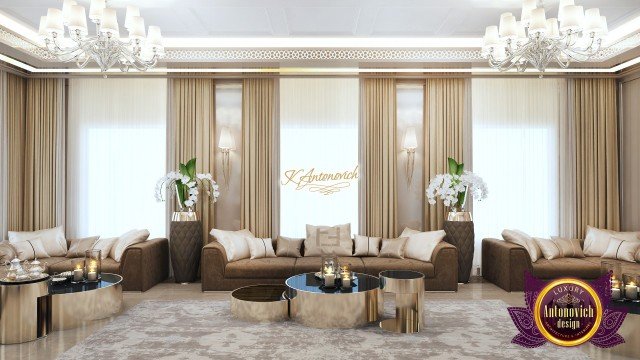 This picture is of a luxurious and modern living room. The room has a cream and white color scheme, with many elements of beige, gold, and silver accents. It has a white couch with two beige armchairs, and a glass and gold coffee table in the center. There is a beige fur rug on the floor, and a large mirror over the fireplace. To the side is a large cabinet with pillows and other decorative items, and tall windows with cream curtains.