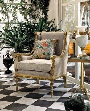 This picture shows a luxurious double-height ceiling foyer with exquisite crystal chandeliers. The walls are made of white marble and feature intricate gold inlays and accents. On the floor is a beige rug that has a raised diamond pattern. Along the edges of the room are high-backed, cream-colored tufted chairs with gold accents around the edges. The large windows let in natural light, and the glass railing allows for an open view.