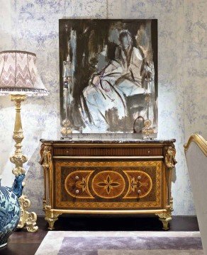 This picture shows a luxurious modern bedroom, featuring a king-size bed with white and gold linens, two marble nightstands with matching lamps, a dark framed chandelier, and a luxurious velvet armchair. The walls are painted in a light grey shade, and a large golden framed mirror hangs above the bed.