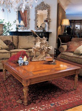 The picture shows a modern living room with luxurious furniture and decor. The walls are painted a warm creamy color and the floor is made of dark hardwood planks. The furniture is an off-white sofa and armchairs, with bright red and yellow embroidered cushions. In front of the sofa is a glass coffee table, and on either side are modern metal and glass shelves filled with books and decorative items. A large vase filled with vibrant flowers stands in the corner, adding a pop of color to the space.