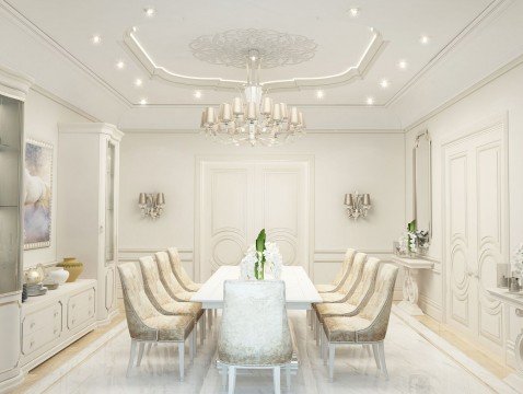 This picture is showing an ornate, modern living room in shades of white, grey, and beige. There are two beige sofas facing each other in the middle of the room, with a white tufted ottoman and a marble-topped coffee table separating them. The walls feature an intricate white wall paneling near the ceiling, adding a luxurious touch to the space. Pops of grey can be seen in the patterned carpet, curtains, and throw pillows.