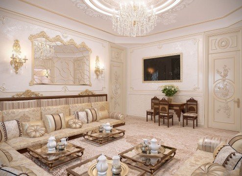 This picture shows a traditional living room with a bright and neutral color palette. The large sectional sofa is upholstered in a pale beige fabric and has plenty of accent pillows in coordinating colors. To the right is a white wingback chair, upholstered in a patterned ivory and beige fabric. On the walls are framed photographs, creating a pleasant visual interest. The floors are finished with a light colored hardwood and an area rug with a subtle pattern. The brightness of the room is further enhanced by two large windows, with white shutters, letting in