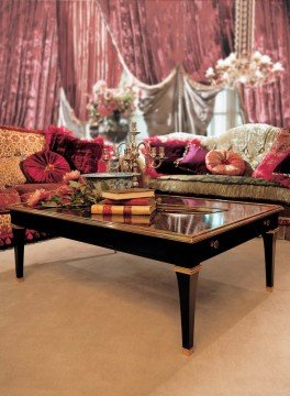 This picture shows a luxurious modern living room with beige walls, bright white and brown furniture, and two large bay windows. The room features two leather armchairs, a L-shaped chocolate-brown sofa, a round glass coffee table, and a wooden bookcase. The floor has a large Persian rug which provides a pop of vivid color and texture to the room. In front of the bay windows, there is a large chaise lounge with white and blue pillows.