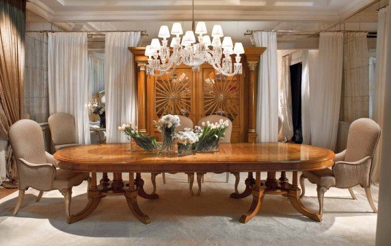 This picture shows a luxurious living room with cream and beige colored walls and furniture. The room includes a large white modern-style sofa, two matching armchairs, a glass and metal coffee table, a decorative area rug, and wall art. The room is decorated to the highest standard with gold accents throughout and elegant chandeliers above.