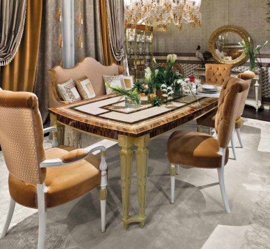 This picture shows a luxurious, contemporary dining room with a long, rectangular marble dining table. The table is surrounded by cream-colored chairs with high backs, and the walls are decorated with elegant wallpaper. On the wall behind the table is a large, golden-framed mirror surrounded by beautiful sconces. Underneath the sconces is a long wooden sideboard, which is decorated with several pieces of art and framed photos. The floor is covered in a plush beige carpet, and the room is finished with a striking crystal chandelier in the center of the ceiling.