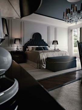 This picture shows a luxurious and modern master bedroom. The room has light gray walls and ceilings, with a unique patterned beige covering the floor. The bed is an elegant white four-poster bed, with decorative bedding and tufted headboard. There is an area at the foot of the bed with two comfortable chairs and a table. On each side of the bed are white nightstands, with matching lamps and wall sconces. On the left side of the room is a large wardrobe built into the wall, with a long mirror above it. The right side