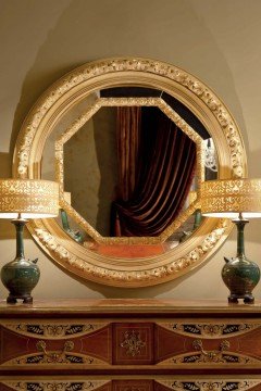 This picture shows a luxurious bedroom with high ceilings and parquet flooring. The walls are decorated with a stunning pattern of light blue and white, and the bed is covered in luxurious velvet bedding. There is a big window to the side, with light pink curtains hanging from the ceiling. To the right there is a round nightstand with a small vase of flowers and a lampshade on top.