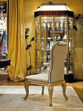 This picture shows a European-style dining room with a large oval table surrounded by matching chairs. The table is covered in a white and gold patterned tablecloth and is topped with a large crystal vase of white roses and greenery. A large chandelier hangs above the table and two ornate paintings hang on opposite walls. A doorway is visible in the background.