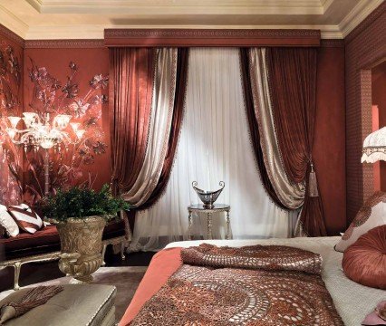 This picture shows a modern-style bedroom with a white upholstered bed and matching chair, both of which have silver nailhead trim. The walls are painted in a light gray color which complements the dark hardwood floor and white curtains. The nightstands are made of a dark wood and feature sleek, black lamps. A large mirror hangs above the bed and a round ottoman sits at the foot of the bed.