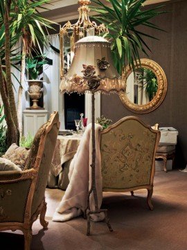 This picture shows a luxury living room with elegant decorations. The walls are a light beige color with contrasting cream and white trim. On the left is an ornate fireplace with a white mantle. There is a large, light brown sofa with several matching ottomans, as well as a glass-top coffee table with matching side tables. To the right is a long, white sofa with several blue pillows. On the walls are several pieces of art, including a large abstract painting, a portrait of a woman, and a cityscape. The floors are made of dark wood,