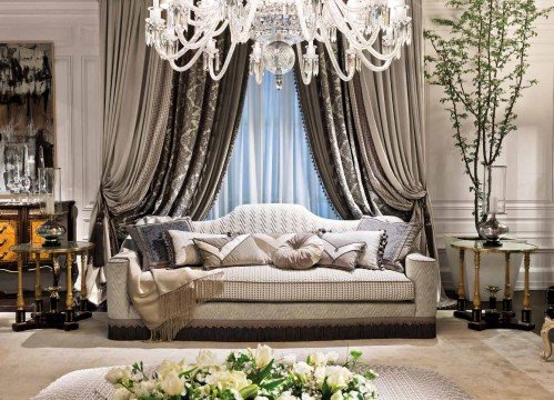 This picture shows an elegant, traditional-style living room with a luxurious white marble floor and walls. In the center is a beige colored sofa with two cream-colored armchairs and a large wooden coffee table with a light grey marble top. On either wall are two golden sconces, with elegant artwork hung between them. There is a large fireplace at the far wall with a white mantel and hearth, and two large double-doors with white curtains draped across them leading onto a balcony.