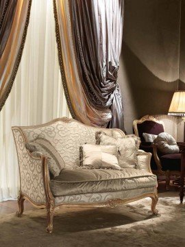 This picture shows an elegant, modern living room space. It features a white and gold accent wall, with a grey and black geometric patterned area rug with hints of pink and white. The sofa is black with white and pink accent pillows and a patterned gold and white throw blanket. There is a round glass coffee table in front of it, alongside two black and gold armchairs with white and pink accent pillows. To the left is a fireplace, with a white marble surround and a white mantel. Finally, there are several plants throughout the room that add to the inviting atmosphere