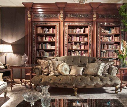 This picture shows a beautiful, modern living room with a large chandelier hanging from the ceiling. The walls are painted in a light grey color and there is large, white built in bookshelf on one side. The floor is covered in a beige marble tile and there is a white sofa and armchair set up in one corner of the room. There is also a small glass coffee table in front of the sofa, and two end tables near it.