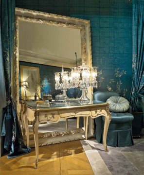 This picture shows a luxuriously decorated dining room. The room features a grand chandelier hanging from the ceiling, ornate wallpaper and curtains, an intricately carved wooden dining table with plush velvet chairs, and an ornamental plant in a large planter.