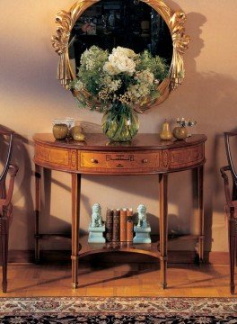 This picture shows an artistic designed living room with a grand wooden chandelier suspended from the ceiling. The room is decorated with a warm, neutral palette of beige and gray colors. There is an ornate patterned rug in the center of the room and two couches featuring gilded accents in the front. A tall indoor plant is next to one of the couches, with a side table located between the two. The walls are decorated with art and framed mirrors.