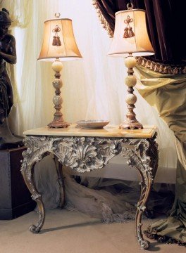 This picture shows a large, ornate and luxurious living room. The walls are covered in an intricate floral pattern, with multiple golden accents and columns throughout. There is a large white marble fireplace and mantle with a black frame. A few pieces of dark wooden furniture are arranged around the room, and an elaborate crystal chandelier hangs down from the ceiling. The whole room is surrounded by floor-to-ceiling windows that offer a view of the outdoors.