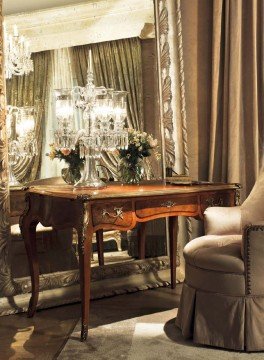 This picture shows an elegantly designed bedroom, featuring a large four-poster bed with draping sheer fabric and a tufted headboard. The bed is centered in the room and is flanked by two night stands. To the right side of the bed is a large armoire, while further away to the left is a lounge chair with a round end table. The walls around the room are decorated with a beige and cream striped wallpaper, accented with a striking patterned area rug and a wall sconce lighting fixture.