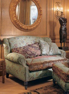 This picture shows a luxury living room designed in a classic style. It features a grand chandelier, a white sofa with an ornate gold frame, an ornate armchair, and two end tables. Other elements include light-colored walls, a large round mirror, and tall windows with bright yellow curtains. Gold accents throughout the room create a balanced yet sophisticated atmosphere.