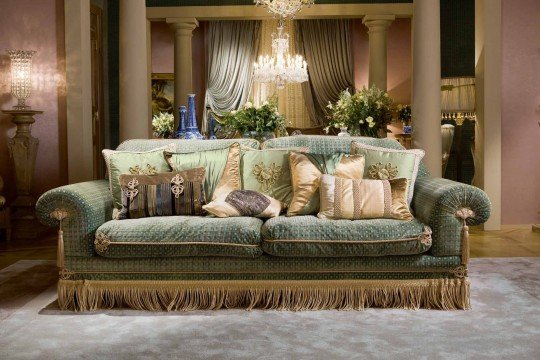 This picture is of an elegant living room space featuring a light blue velvet sofa, two armchairs upholstered in a patterned fabric, and a glass-topped coffee table. There is a white fireplace with a cream mantlepiece and a round blue mirror above the fireplace. On either side of the fireplace are two glass display cabinets filled with books and decorative objects. The floor is a light hardwood with a white and grey patterned rug underneath the seating area. The walls are painted in a pale blue tone to match the sofa and there is a large window allowing plenty of natural