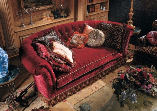 The picture shows a creamy white interior room, with a woman sitting on a navy blue sofa. The walls are painted in a light grey colour, and there is a round glass coffee table in the middle of the room. To the right, there is a fireplace with a large mirror above it. On the walls are several geometric paintings that add a vibrant pop of colour to the room.