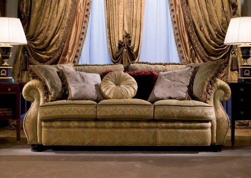 This picture shows a luxurious dining room in an opulent home. The room features sumptuous white walls with ornate moldings on the upper walls and doors, a dark wood floor, and rich velvet curtains hanging from the windows. There is an elegant dining table set for a meal, with white and gold plates, crystal goblets, and silverware, as well as a crystal chandelier above it. Along the back wall is a beautiful sideboard with a display of flowers, a mirror, and other décor pieces.