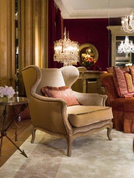 This picture shows a luxurious living room, featuring a beautiful and modern beige sectional sofa and armchairs in front of a white marble fireplace with a wood mantel. The walls are covered in a luxurious brown velvet fabric and the space is also accented with two silver chandeliers and two round accent tables. On either side of the fireplace are two tall vases filled with lush green plants.