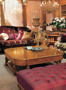 The picture shows an intricately decorated living room. It features a black grand piano, a light-colored marble fireplace with wooden mantle, and two black couches. The walls are covered in a cream-colored wallpaper with a floral pattern, and the furnishings have a touch of gold. A beige rug pulls the look together, and two sets of drapery frame the windows.