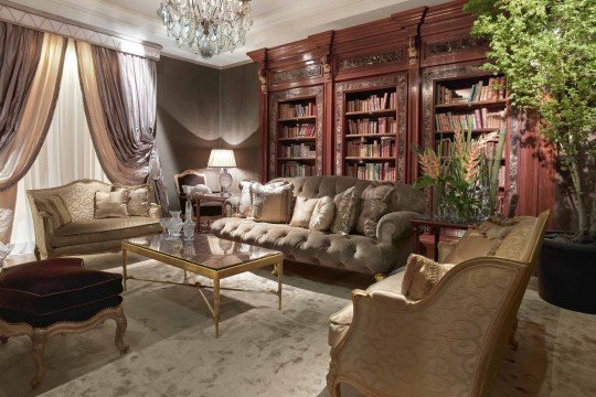 This picture shows an interior with an elegant design. The walls are covered in a blue and silver geometric wallpaper pattern, and in the center of the room there is a tufted blue velvet couch with gold accents. On the right side of the picture is a white marble fireplace with a flat-screen TV mounted above it. There are also two white chairs and a glass coffee table near the couch, and a large plants on the wall behind the furniture.