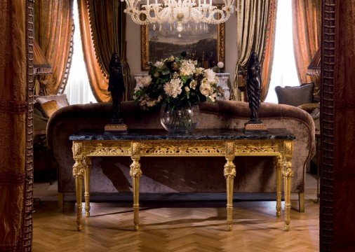 This picture shows a luxurious bathroom with intricate gold detailing and marble accents. The bathroom includes a large bathtub, a rain shower, a marble vanity with two sinks, and a separate area with a toilet. The walls are a pale beige color, while the floors are made of black and white mosaic tiles. Gold accents can be seen in the light fixtures, the faucets, and various decorative elements throughout the room.