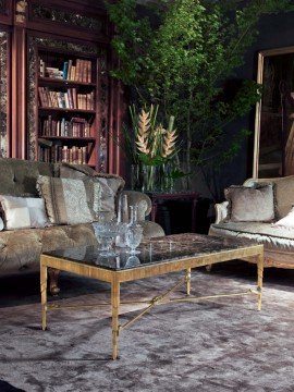 This picture shows a luxury, modern living room. The room has white walls and dark wood floors, and the furniture includes a comfortable white sofa, a couple of wooden armchairs, and a round glass-top coffee table. There are also a few colorful throw pillows and a large, ornate mirror hung on a wall. The room is lit by several stylish ceiling lights and tall floor lamps, and an attractive area rug finishes off the look.