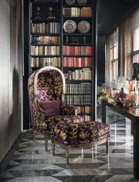 The picture shows the interior of a large, luxurious house. It features an ornate marble and wood staircase, along with a crystal chandelier hanging from the ceiling and framed artwork adorning the walls. The floor is covered in a luxurious red and gray patterned rug. There is also a modern blue velvet sofa situated at the bottom of the stairs.