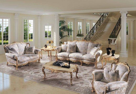 This picture shows a modern living room with clean lines and contemporary furnishings. The walls are painted a light gray color and accented with dark gray moldings. The floor is covered with a textured beige carpet, and there is a single large window in the center of the wall. A black leather L-shaped sectional sofa sits on one side of the room, while two matching armchairs sit at an angle to each other on the opposite side. A square glass coffee table sits in front of the sectional, and an ornate cream-colored console table stands against the wall