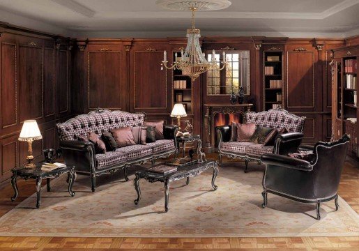 This picture shows a luxurious living room with vaulted ceilings and a formal seating area. The walls are painted a deep burgundy color while the furniture is a combination of white and a muted gold. There is a large crystal chandelier in the center of the room and two wall sconces lighting up the space. The floor is tiled in black and white checkerboard pattern and an area rug in a complementary shade of burgundy adds a touch of warmth.