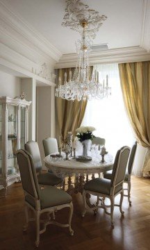 This picture shows an elegant and luxurious living room interior. It features a grand, light-colored sofa set with ornate detailing, two white chairs with tufted cushions, a low table with intricate legs, and a statement rug in dark hues. There is a large framed artwork piece on the far wall, and the room also has a pair of matching end tables, drapes and curtains, and a sideboard with glass doors.