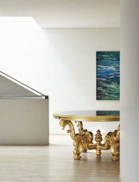 This picture shows a luxury living room decorated in a modern style. The walls are painted a pale shade of grey, and the floor is covered in a light-colored carpet. The furniture includes a low-set white couch and two matching armchairs upholstered in blue fabric. A white coffee table sits in between them and the center of the room features a modern crystal chandelier. In addition, there are several pieces of artwork hanging on the walls and two small tables with lamps beside the couch.