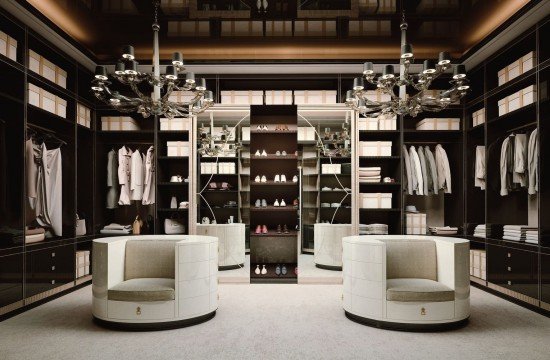 This picture shows a modern, luxurious living room. The walls are an off-white color with an ornate archway and tall windows that provide natural light. There is a gray sofa against one wall decorated with grey and white throw pillows, while in the corner there is a black and silver chandelier. A large rug covers most of the floor space, and off to the side there is a white fireplace. There is also a black bar cart with glass shelves, which adds an elegant touch.