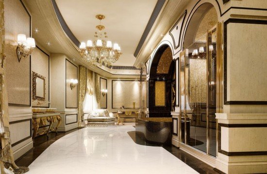 This picture shows an ornate double staircase leading up to a beautiful, grand entrance hall. The walls are covered in luxurious white marble, and the ceiling is intricately patterned with golden designs. The stairs have white, gold, and red balustrades and handrails, which lead up to a gated landing in front of two large, intricately carved wooden doors. A large chandelier hangs from the center of the ceiling, giving the room an air of opulence and luxury.