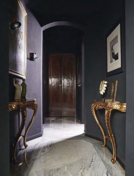 The picture shows an elegantly designed modern foyer. It features a marble flooring with a geometric pattern, a tall ceiling with intricate designs, and two round pillars with gold accents. The walls are finished with luxurious paint, and a large chandelier hangs from the ceiling. The foyer also has a large rectangular table with two chairs, and a mirror adds the finishing touch.