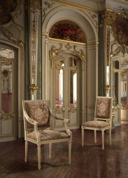 This picture shows a luxurious, modern living room. It features an ornate dark velvet sofa with two large tufted cushions, as well as a matching armchair. The room is decorated with a white and gold patterned rug, a large ornate mirror, and two ornately carved wooden side tables. Artwork is displayed on the walls. The room also features a large chandelier, and its windows are covered by heavy velvet curtains.