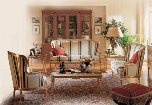 This picture shows a large, spacious living room with classical interior design elements. At the center of the room is a silk sofa with an off-white and gold floral pattern. The walls are painted in a warm tan color and decorated with intricate wood paneling. The ceiling has a decorative wooden beam, and two grand chandeliers hang from it. The room is filled with classic furniture pieces, such as an armchair, two occasional chairs, and a side table. Lush green plants in tall vases add a natural touch to the space.