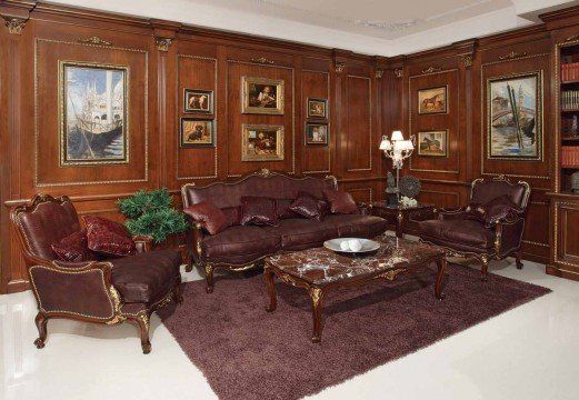 This picture shows a luxurious, contemporary living room with terracotta-colored walls and a high, vaulted ceiling with skylights. The room features a plush, white sofa with ornate gold detailing, two grey armchairs, and a decorative rug with a beige and blue pattern. On the wall above the sofa is an eye-catching piece of art featuring an abstract teal design. A round glass coffee table sits in front of the sofa and there are two wooden side tables with lamps. There are also several potted plants scattered around the room, adding a touch of gre