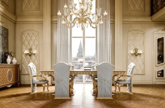 This picture shows a luxurious bathroom with marble floors, walls and countertops. The countertop has a large white double sink with gold fixtures, and there is a separate shower area with a glass door. There is also a large bathtub with a tiled deck for seating, as well as a window with an ornate treatment above it. The walls are decorated with framed artwork, and the room is lit with recessed lighting.