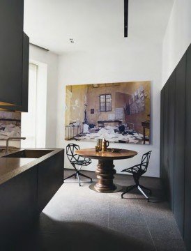 This picture shows a beautiful indoor living area with contemporary furniture, decor and design. The walls are painted in a light gray color and feature multiple art pieces hung on them. The room is well lit with natural lighting, coming through the large windows with sheer drapes. In the center of the room is a modern round coffee table with minimalistic white seating and colorful accent pillows. A patterned rug lies beneath the seating, adding another visual element to the space.