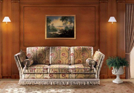 This picture shows a luxurious bedroom with a grey velvet canopy bed and a tufted indigo blue velvet headboard. On the bed are white, grey and light pink bedding, with a stylish ornate mirrored nightstand nearby. There is a white fur rug on the floor and a large crystal glass hanging ceiling chandelier. The curtains are velvet and hang from gold rails, and the grey wall behind the bed is filled with art frames.