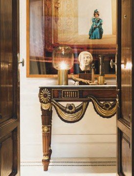 The picture shows a young girl standing in front of a mirror, looking into the reflection of herself. She has two different colored ribbons in her hair and is wearing a blue dress. Behind her, there are shelves with various items on them.