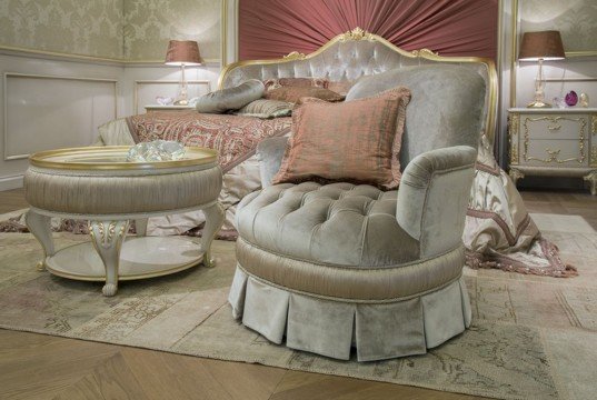 This picture shows a luxurious bedroom with wooden paneled walls featuring intricate gold decorations and a mirrored door decorated with beige silk velvet. There is a large four-poster bed with a lightly patterned duvet cover and an ornate headboard. There is a round accent table on either side of the bed with gold lamps, and a chaise lounge topped with an array of fluffy cushions in the corner of the room. A small side table with a large white flower arrangement sits beside the chaise. The room has a rich burgundy colored rug and a few artworks hung on