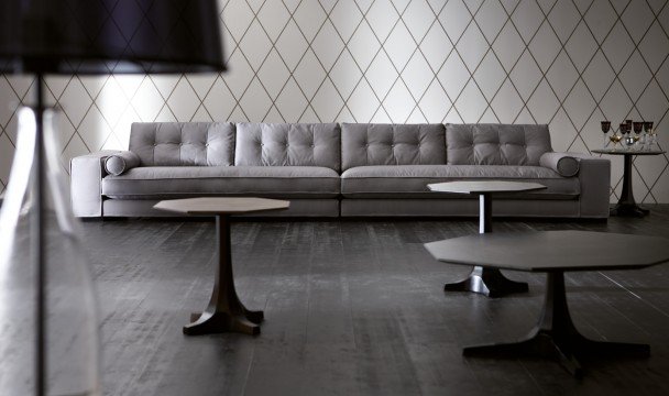 This picture shows the interior of a modern and luxurious living room. There are two beige sofas placed in an L-shape around a white coffee table, with a large cream rug underneath. On the walls of the room, there is a light wooden paneling that extends up to the ceiling and is filled with golden frames holding elegant artwork. To one side of the room, there is a small but stylish bar with mirrored panels and wine glasses arranged on the shelves. On the opposite wall, there is a glass display case full of delicate china. The overall look is one of