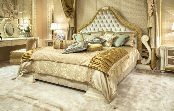 This is a picture of a luxurious living room. The room features an elegant beige and gold interior design with plush furnishings. There is a large white leather sofa, two gold armchairs, a glass coffee table and an ornate antique-style chandelier. On the walls, there are patterned golden wallpaper, paintings and mounted mirrors, along with soft lighting in the corners of the room. The ceiling is decorated with intricate designs and the floor has an area rug with a floral pattern.