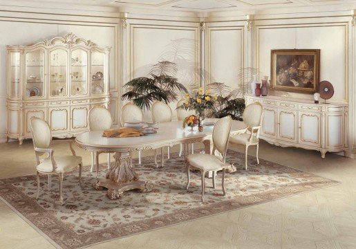 This picture shows a large, luxurious bedroom with an ornate decor scheme. The walls are a soft, creamy white, and the floor is covered with a plush beige rug. The bed has a cream-colored headboard, velvet bedding, and throw pillows of various shades of gold. Two elegant armchairs with gold upholstery, a side table, and a large wall mirror complete the look. Overall, this is a sophisticated and inviting space.